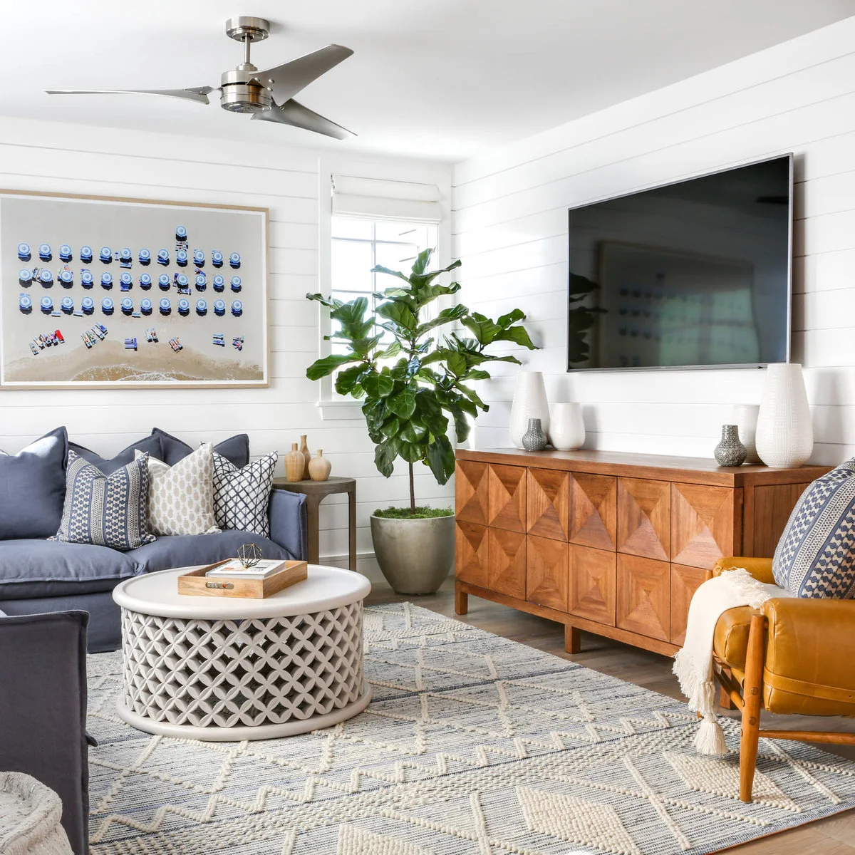 White and small gray vessels sit on a geometric-front wood cabinet below a wall-mounted television beside a leafy floor plant.