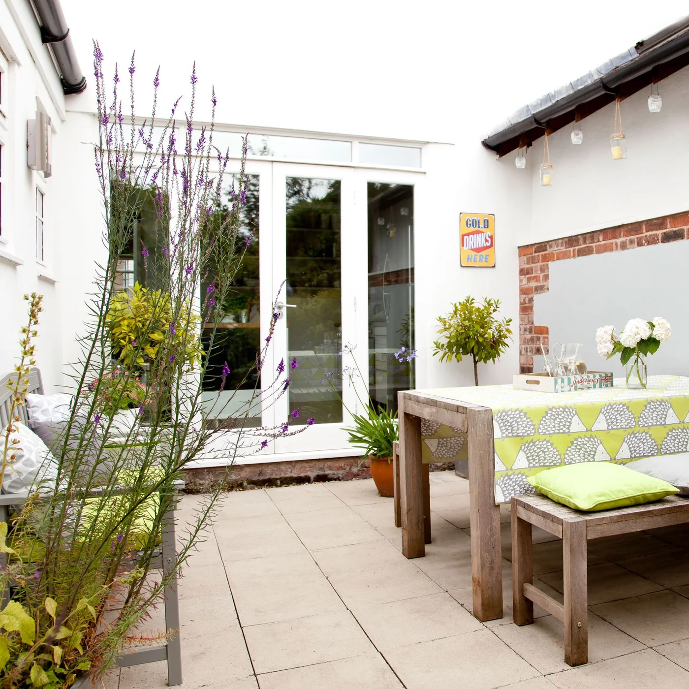 A timber table and bench sit in a pale courtyard with glazed doors and a narrow flowering planter