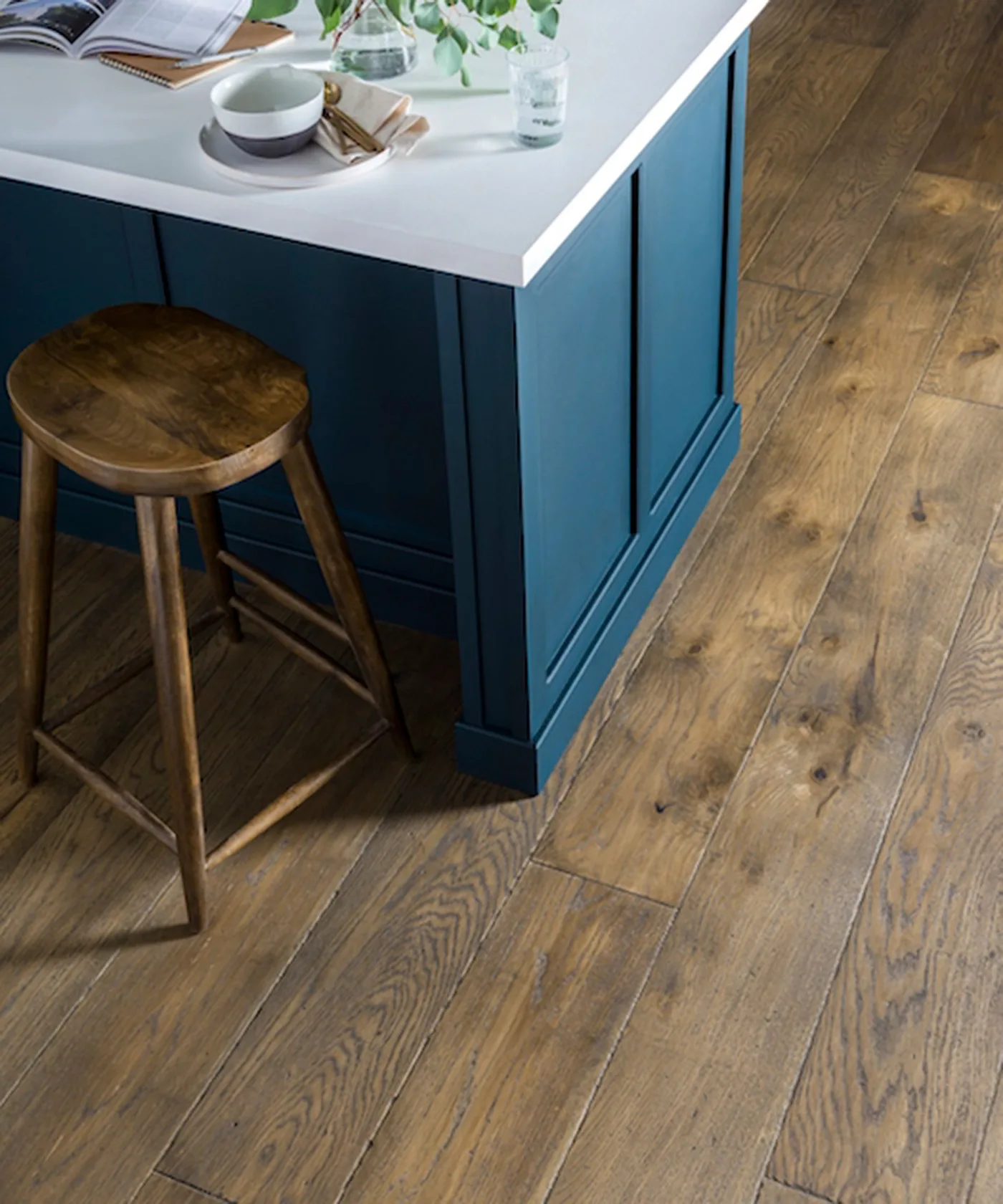Close view of deep blue paneled kitchen island with white counter, wooden round stool, wood floor, cup, books, and small plant.