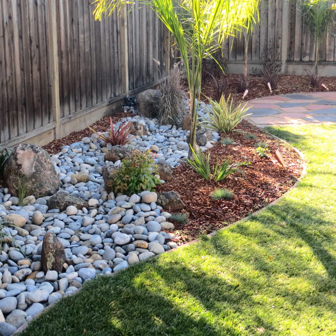 A curving bed of pale rounded stones sits beside bark mulch, small plants and a wooden fence