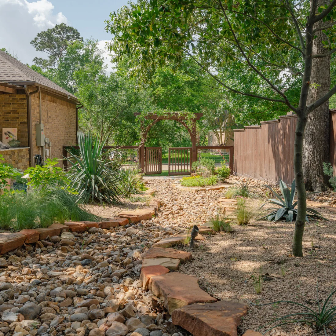 A long river-rock channel with flat reddish edging leads toward a wooden garden gate