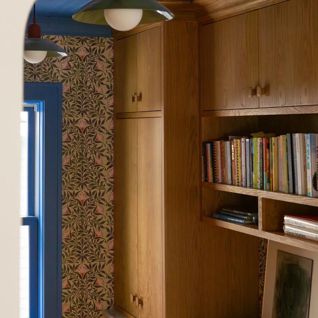 Plain wood entry cupboards and open book niches stand beside leaf-patterned wallpaper and a blue door frame beneath simple pendants.